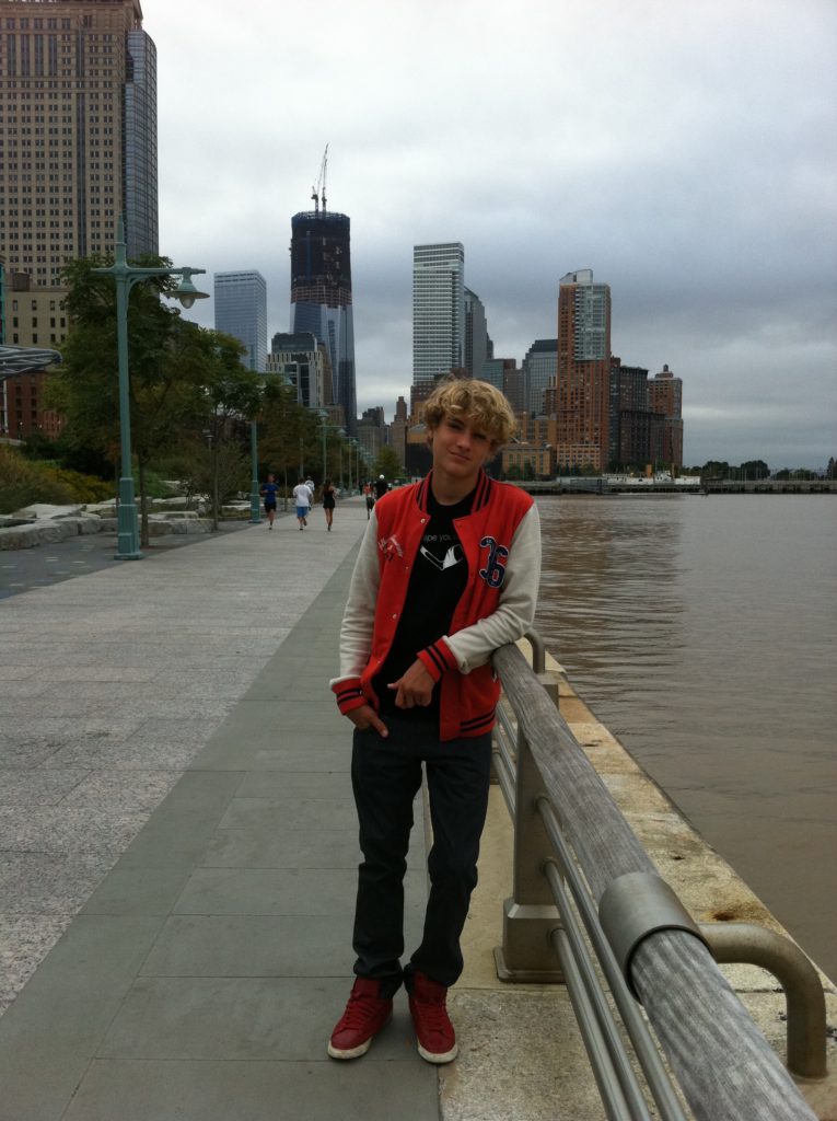 A teenage boy in an orange vest and red sneakers standing along the West Side Highway in downtown Manhattan.