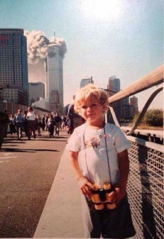 A young child is standing on a street with a pair of yellow binoculars around his neck. Behind him, one of the World Trade Center towers is smoking. The people behind him appear calm.