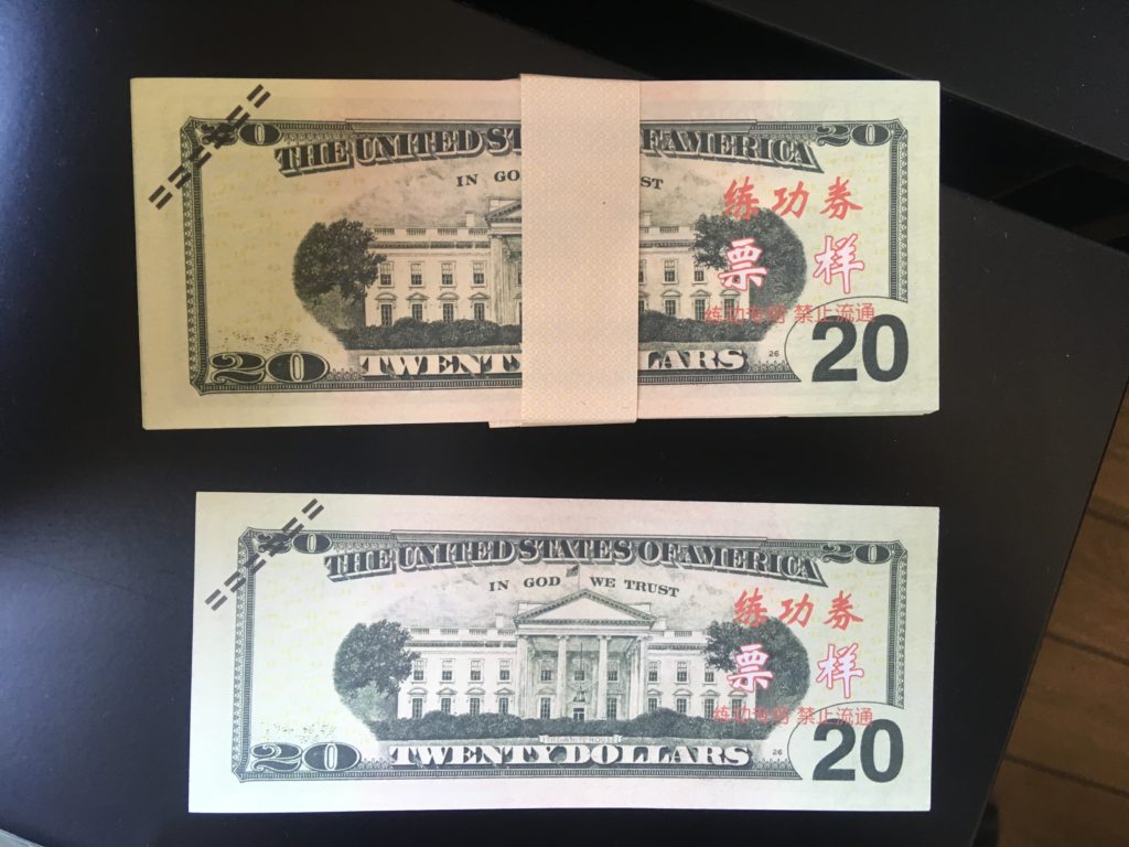 Stack of $20 bills in a paper belt, next to a single $20 bill. Both have red Chinese writing and dashed black lines in the upper left corner