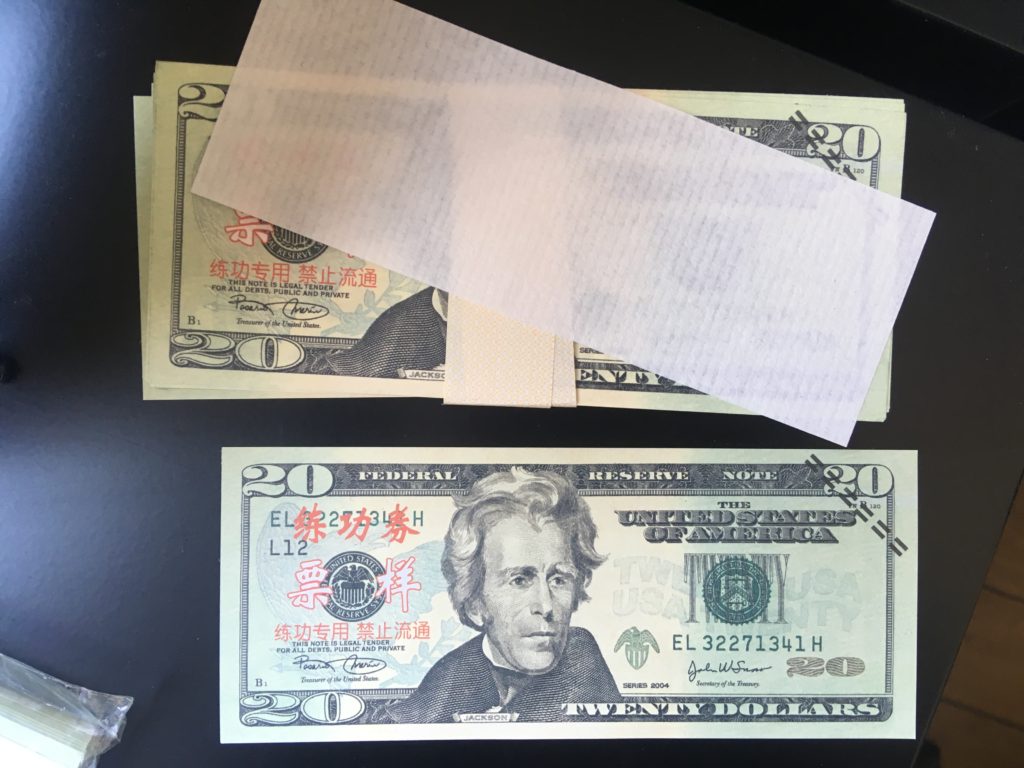 A stack of $20 bills with red Chinese writing partially covered by a piece of paper, wrapped in a paper belt, next to a single $20 bill with Chinese writing.